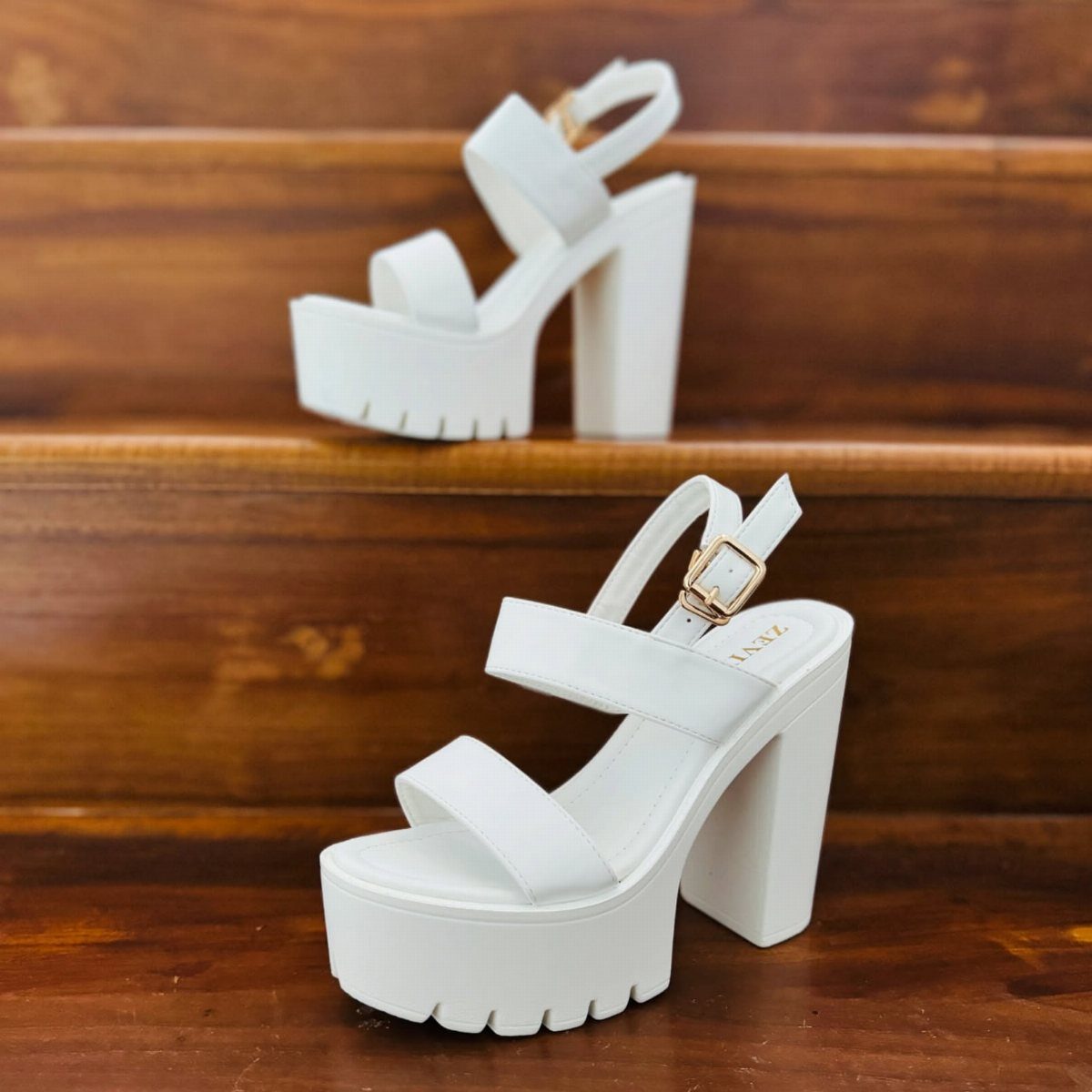 Two white chunky heel platform sandals with wide straps sitting on wooden stairs.