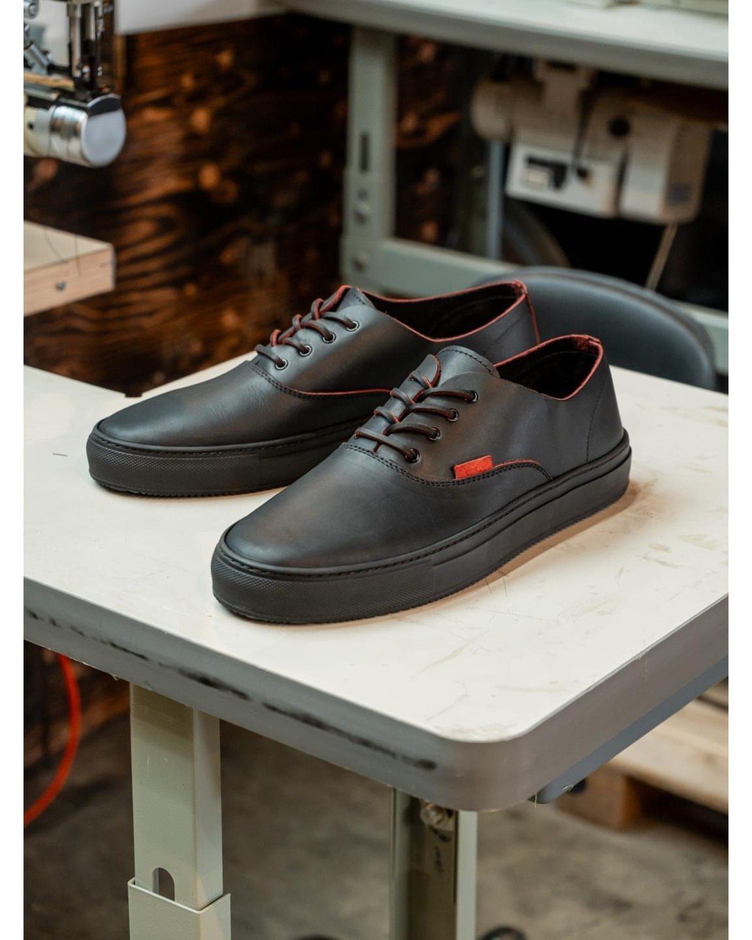 Black leather low-top sneakers with red laces sitting on a white workbench in a workshop.