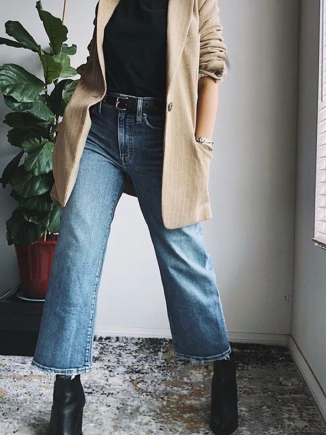 Person wearing blue cropped wide-leg jeans, a tan blazer, and black ankle boots standing near a plant.