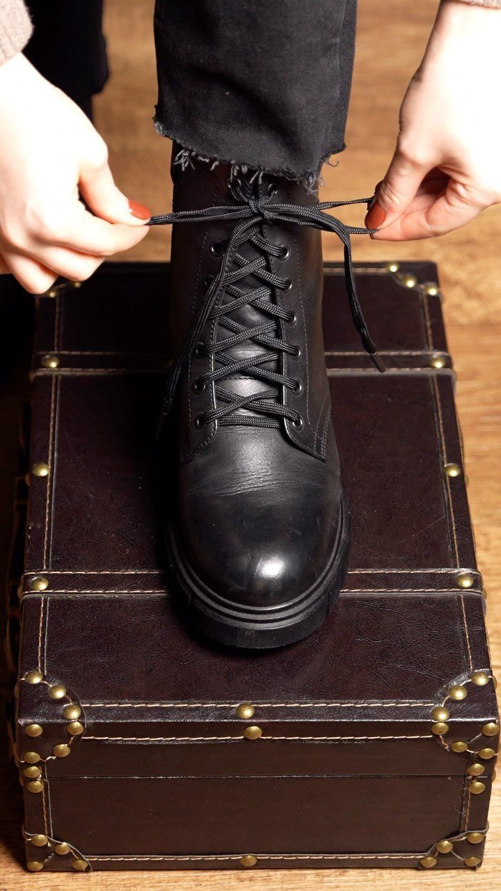Hands tying the black laces of a leather ankle boot worn with black jeans.