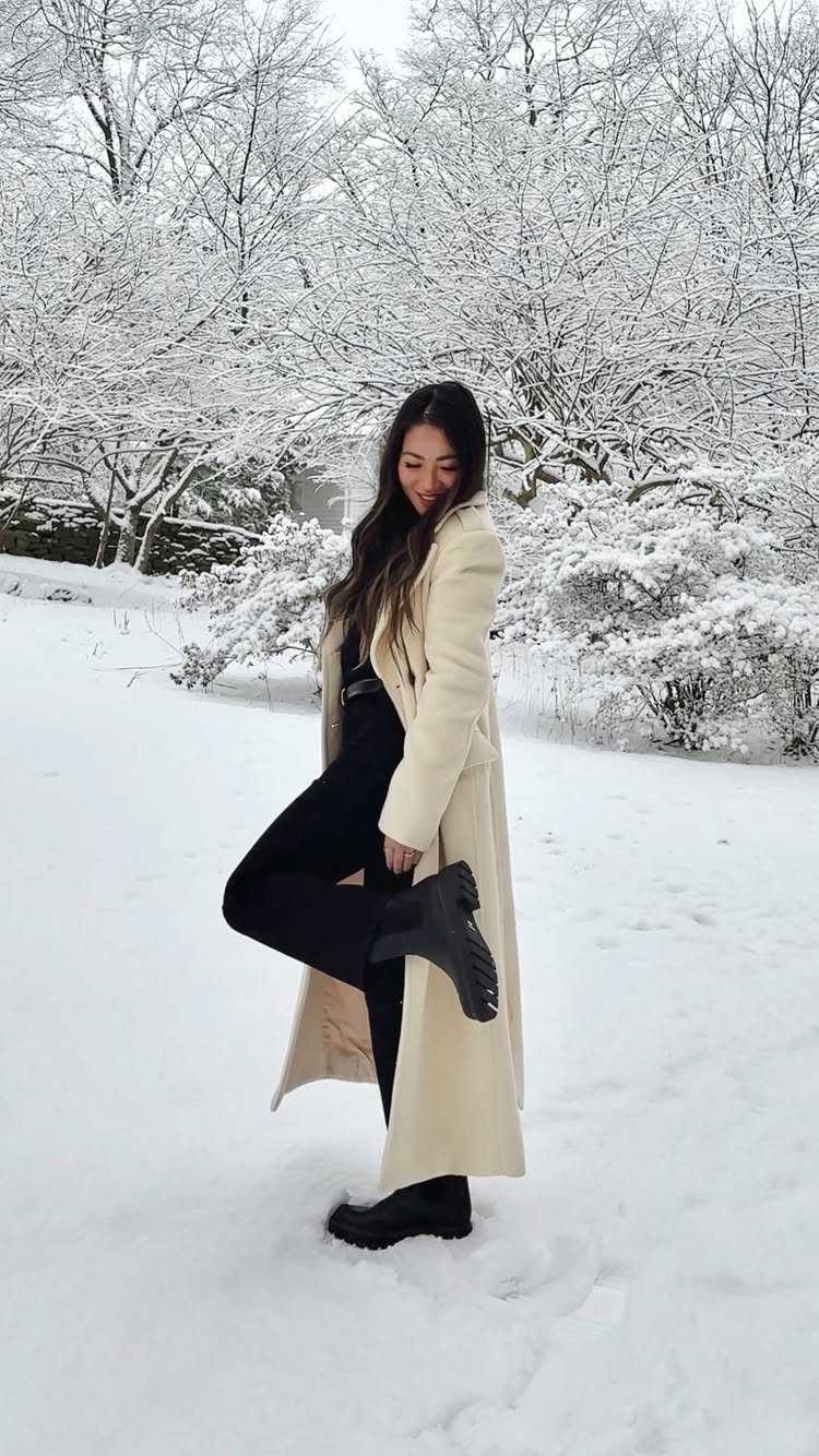 A woman wearing a cream coat and black pants poses in the snow, showcasing her black lug sole boots.