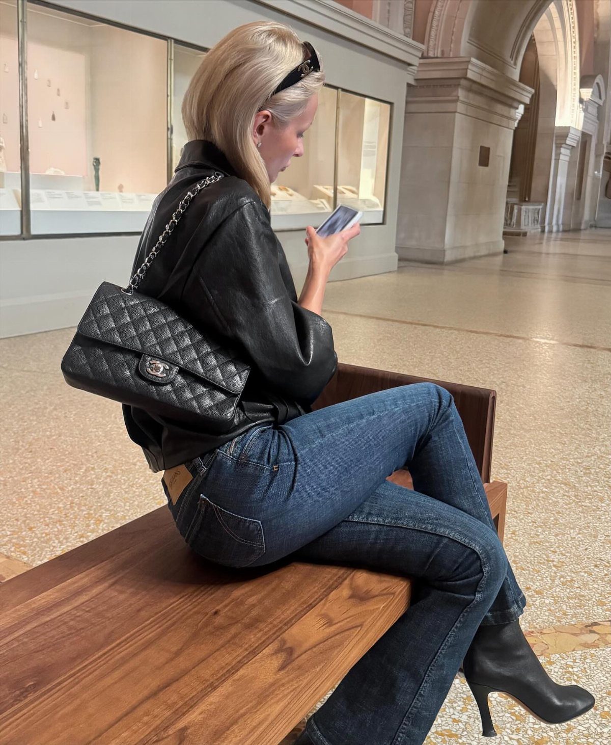 A woman sitting on a wooden bench wearing blue flared jeans and black pointed-toe boots.