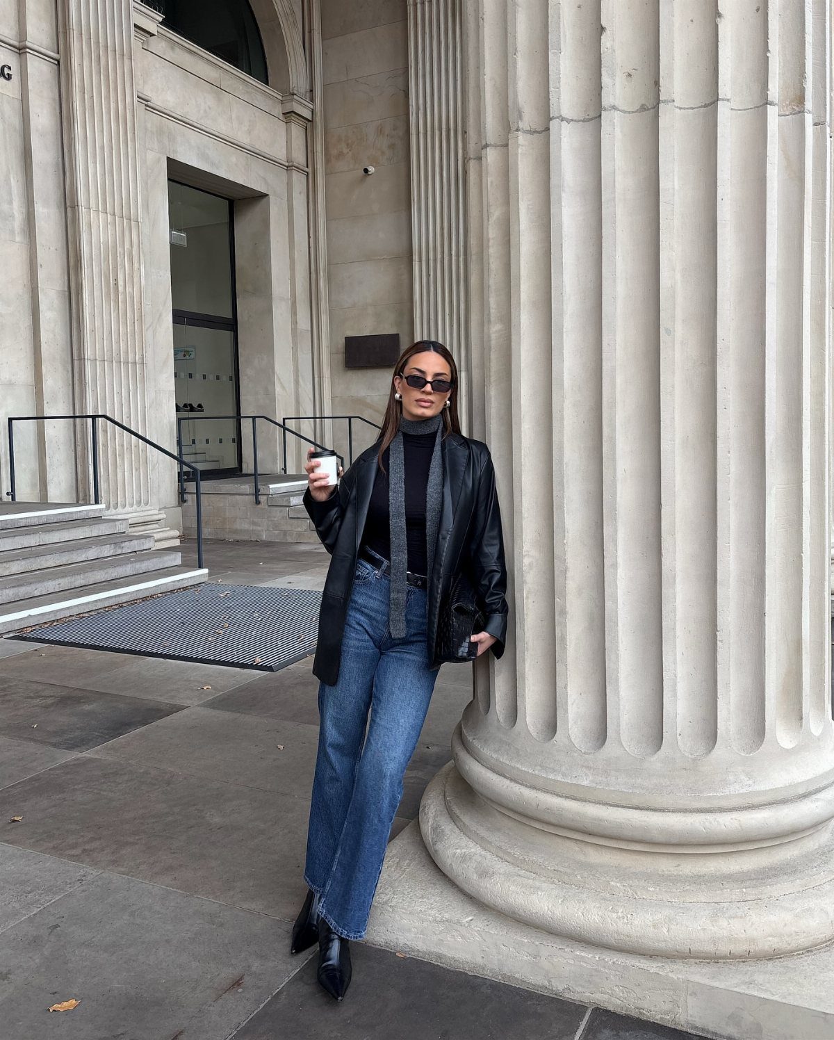 A woman in a leather jacket and blue straight-leg jeans posing by a large column.