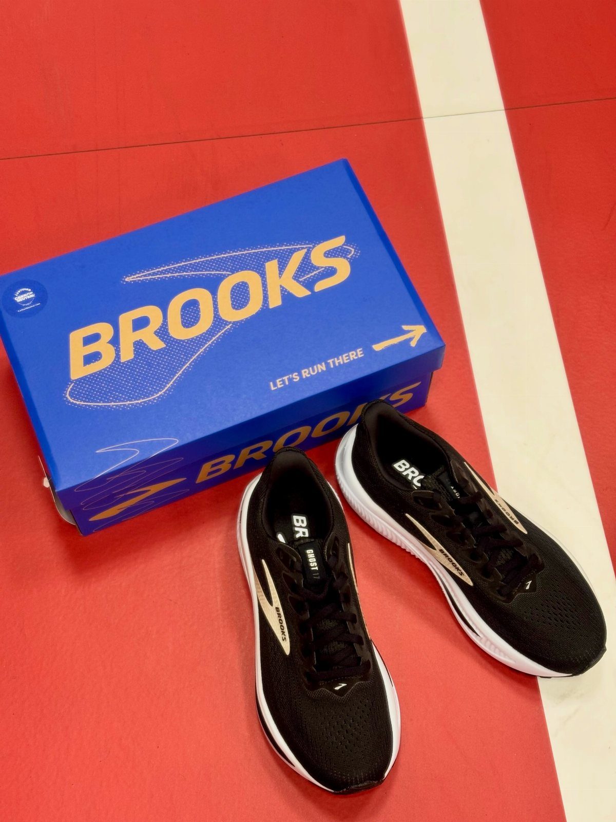 Black Brooks athletic shoes resting beside a blue shoebox on a red and white floor.