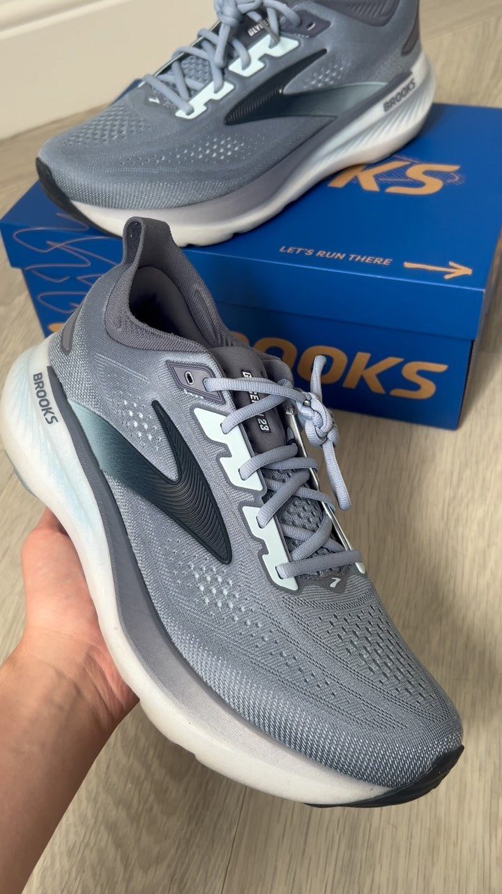 A hand holds a grey Brooks Glycerin 23 athletic shoe above its blue shoebox.