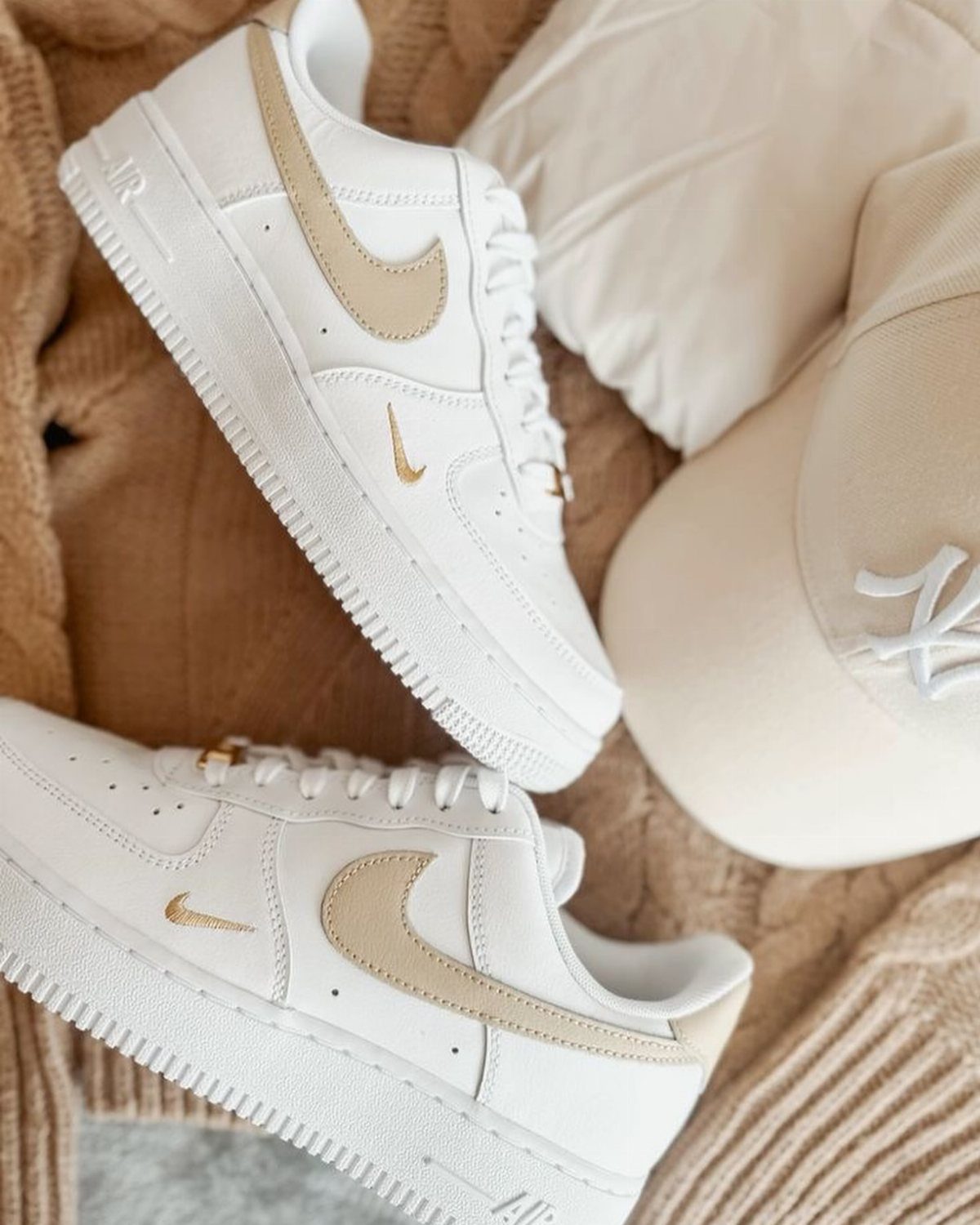 White Nike Air Force 1 sneakers with beige swooshes resting on a tan knit blanket.