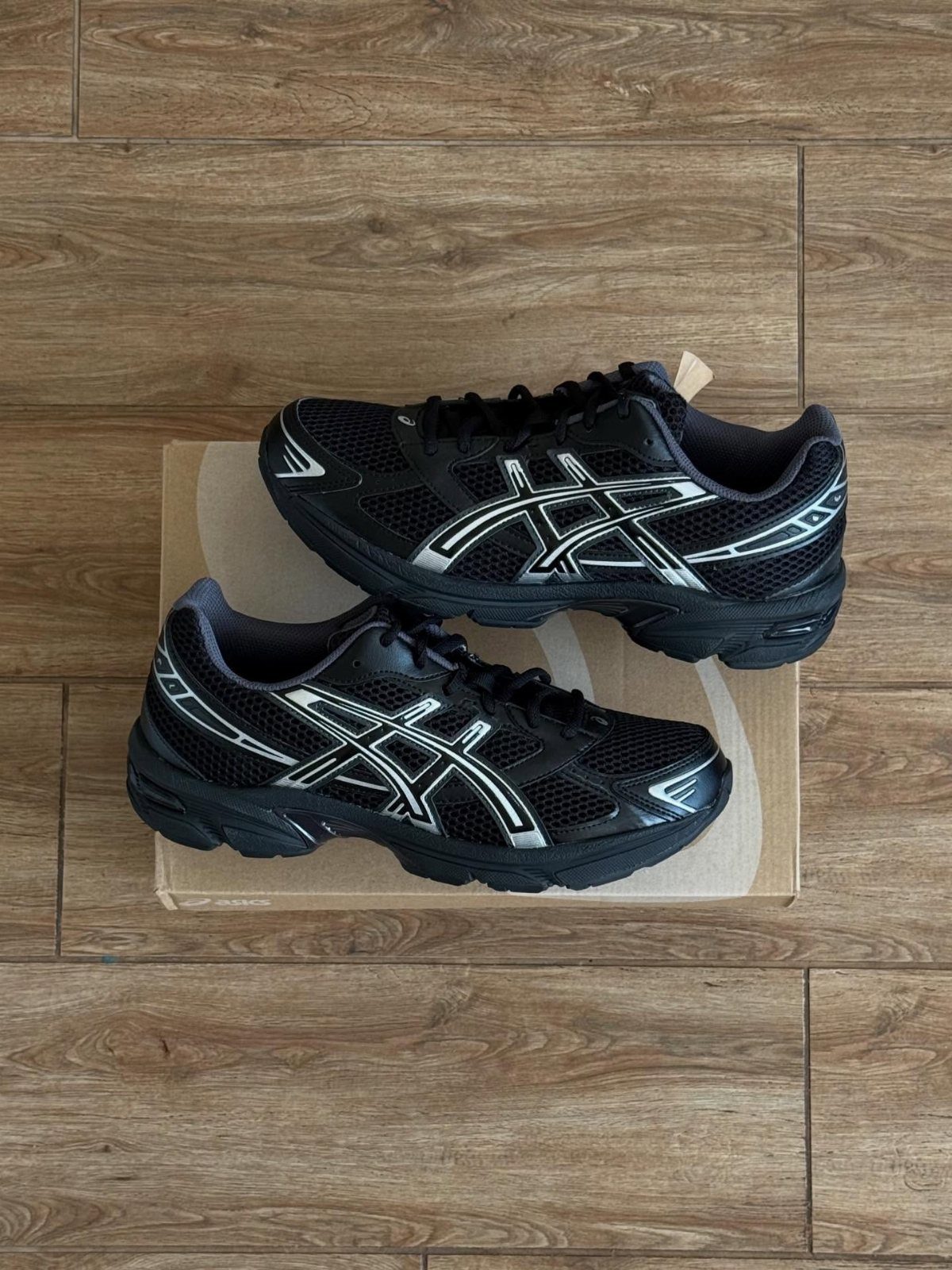 Black and silver ASICS GEL-1130 sneakers sitting on a cardboard box on wooden flooring.