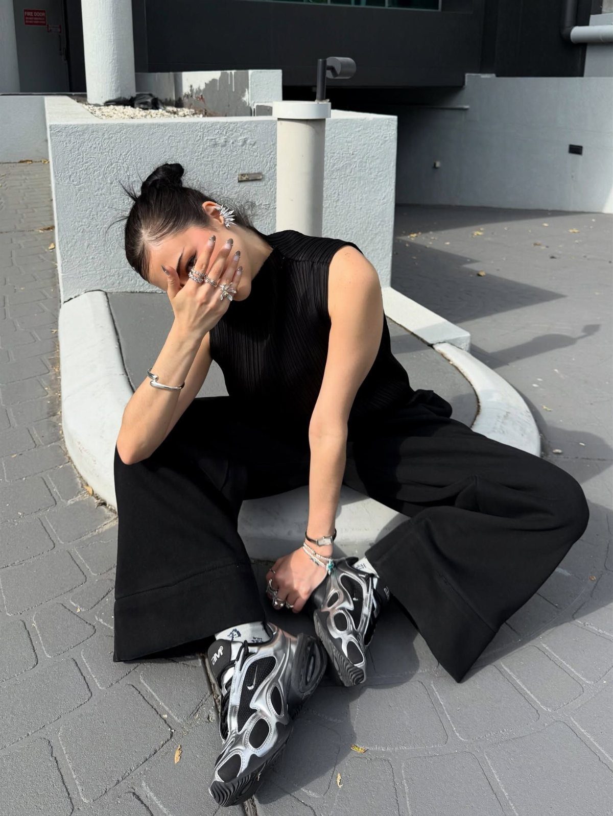 A person sitting outdoors wearing black wide-leg trousers and black and silver sneakers.