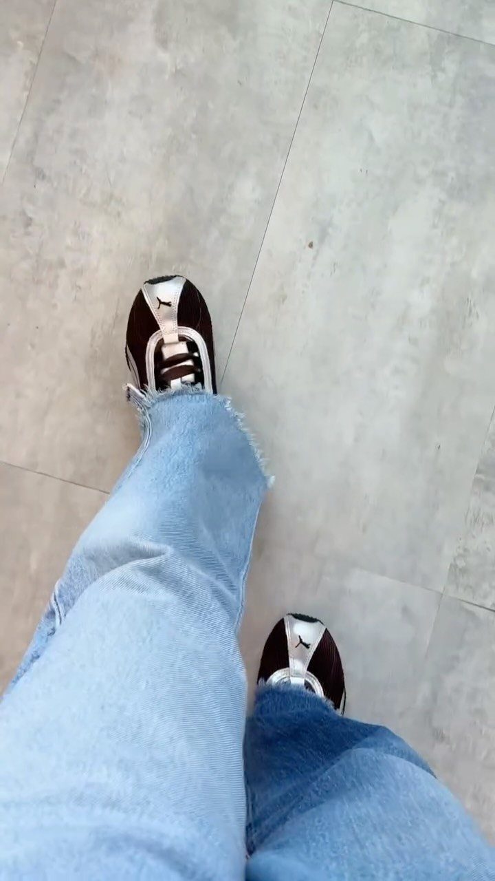 Person wearing light blue jeans and black and silver sneakers standing on grey tiles.