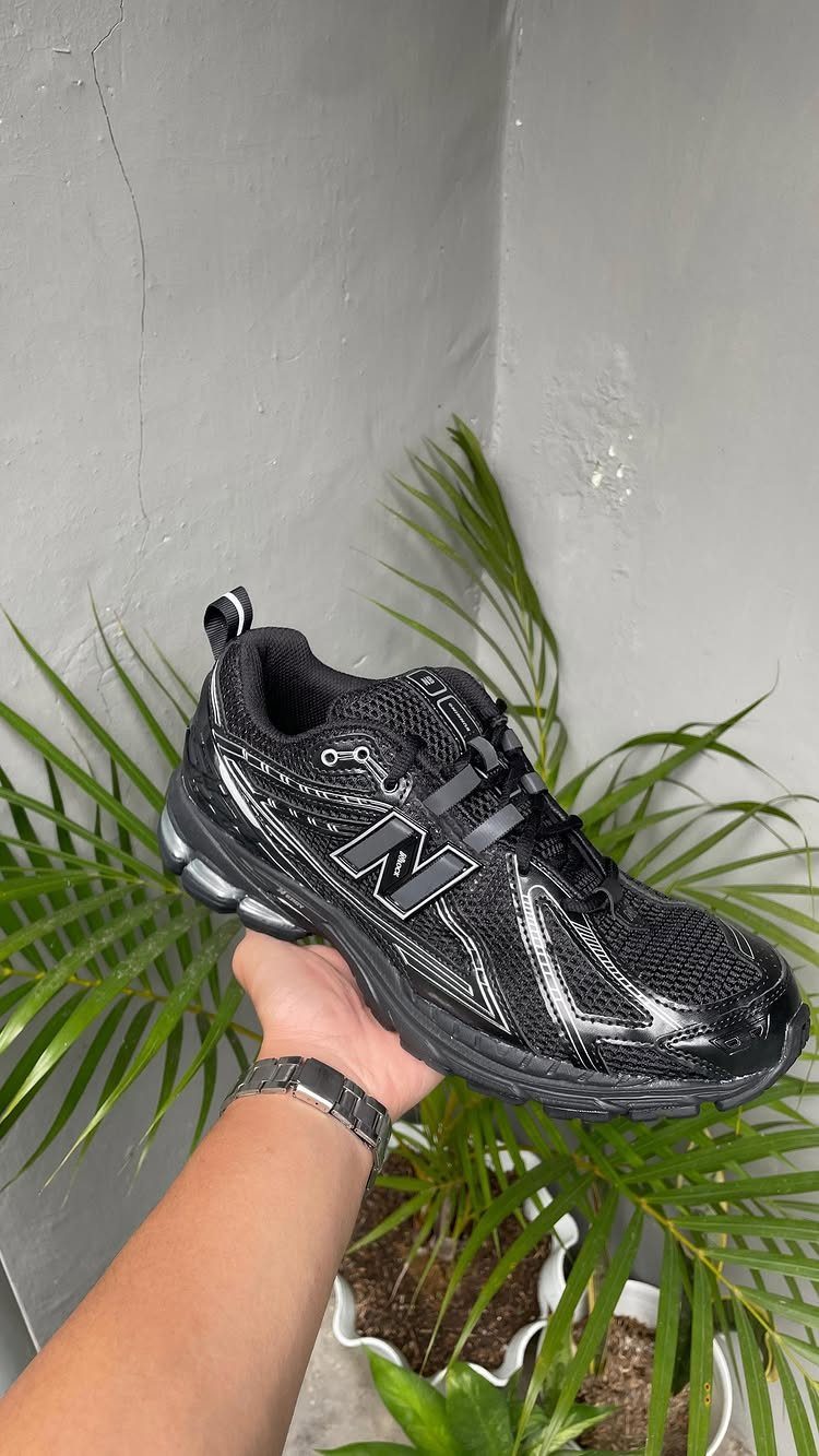 A hand holding a black and silver New Balance sneaker against green palm leaves.