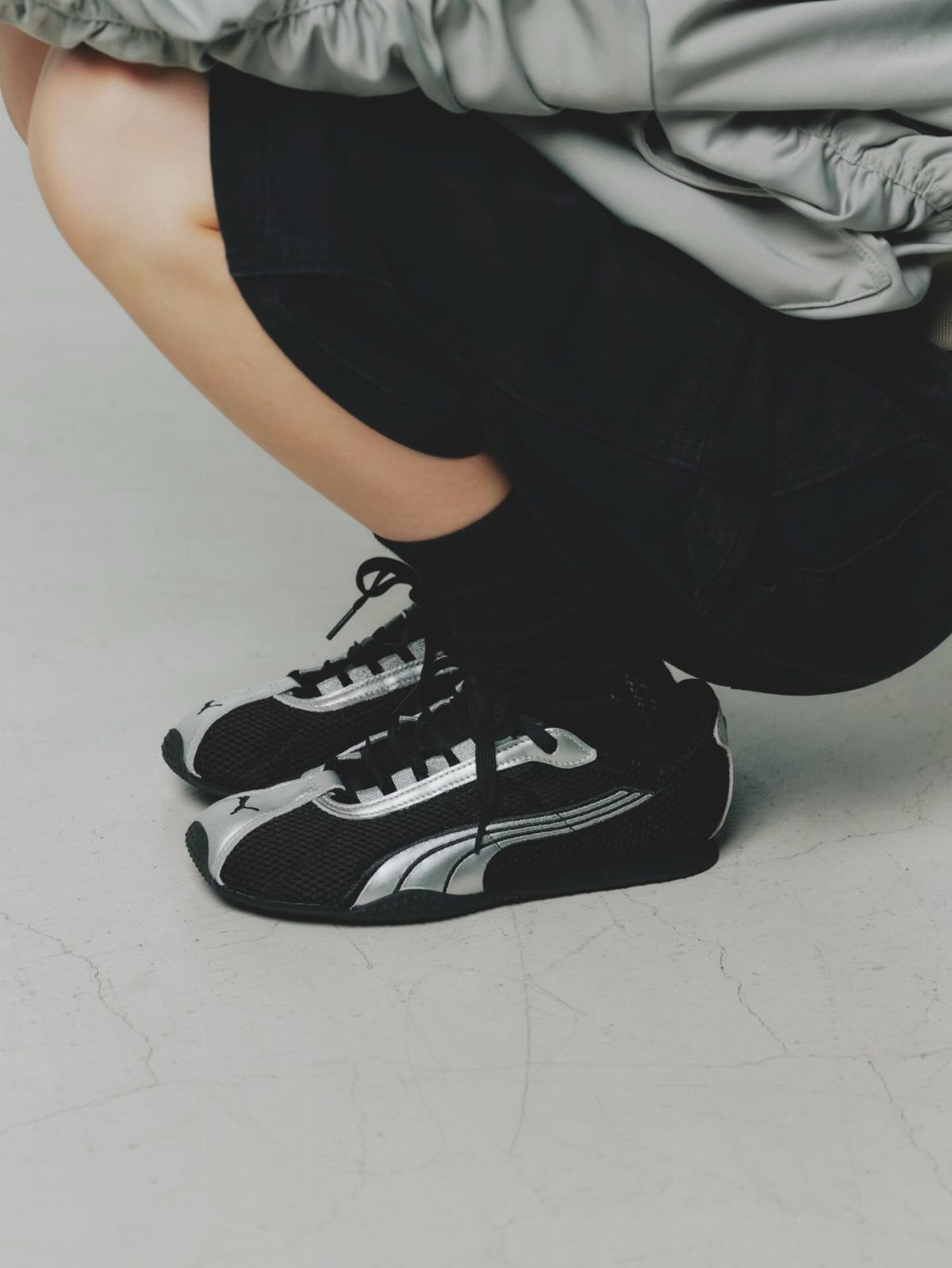 Black and silver PUMA sneakers worn by a person crouching on a light grey floor.