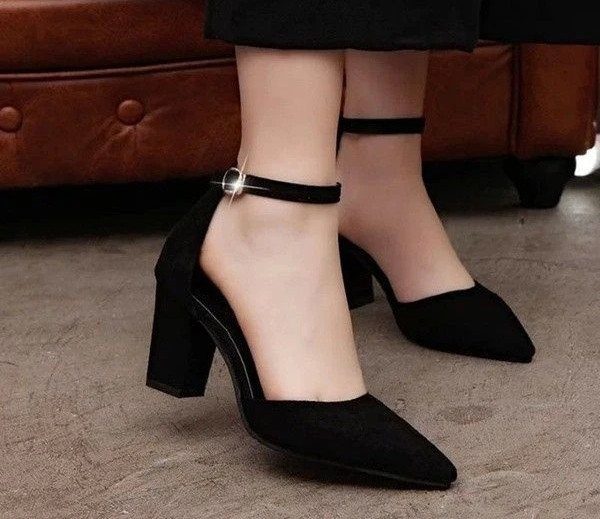 Black velvet pointed-toe pumps with ankle straps worn by a person standing near a leather sofa.