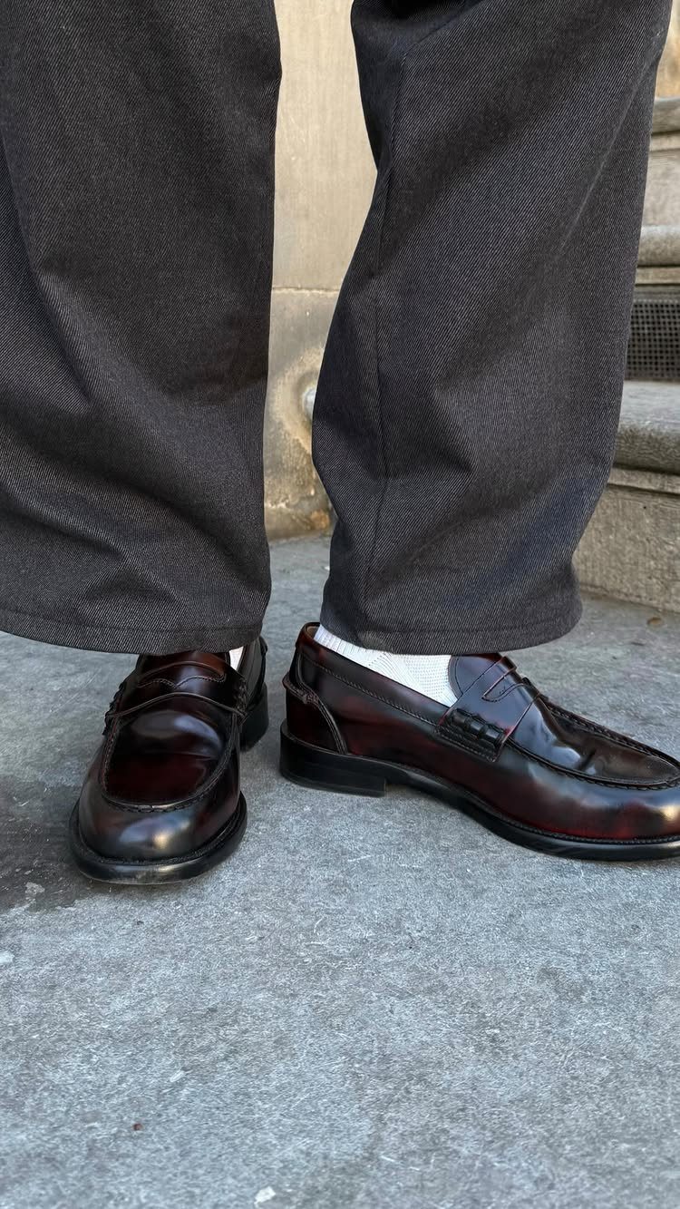 Black patent leather loafers with red soles worn with grey trousers and white socks.