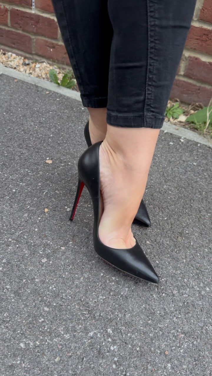 Person wearing black pointed-toe high heel pumps with red soles and black jeans on pavement.