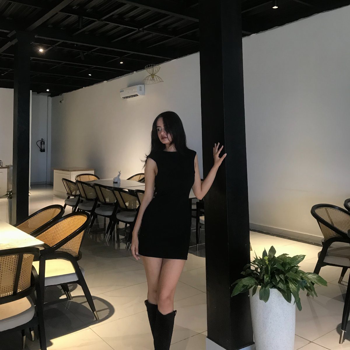 A woman in a black mini dress and tall black boots leaning against a pillar.