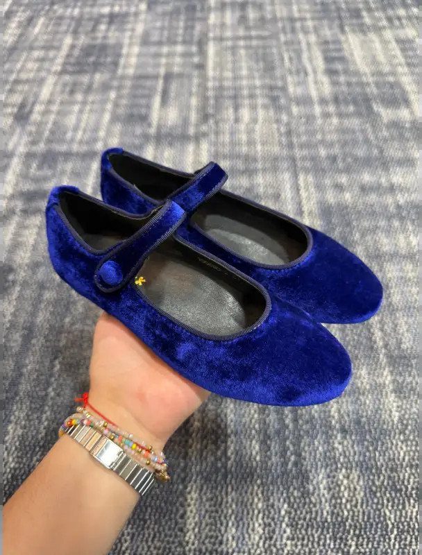 Cobalt blue velvet Mary Jane flats held by a person's hand against a grey textured background.