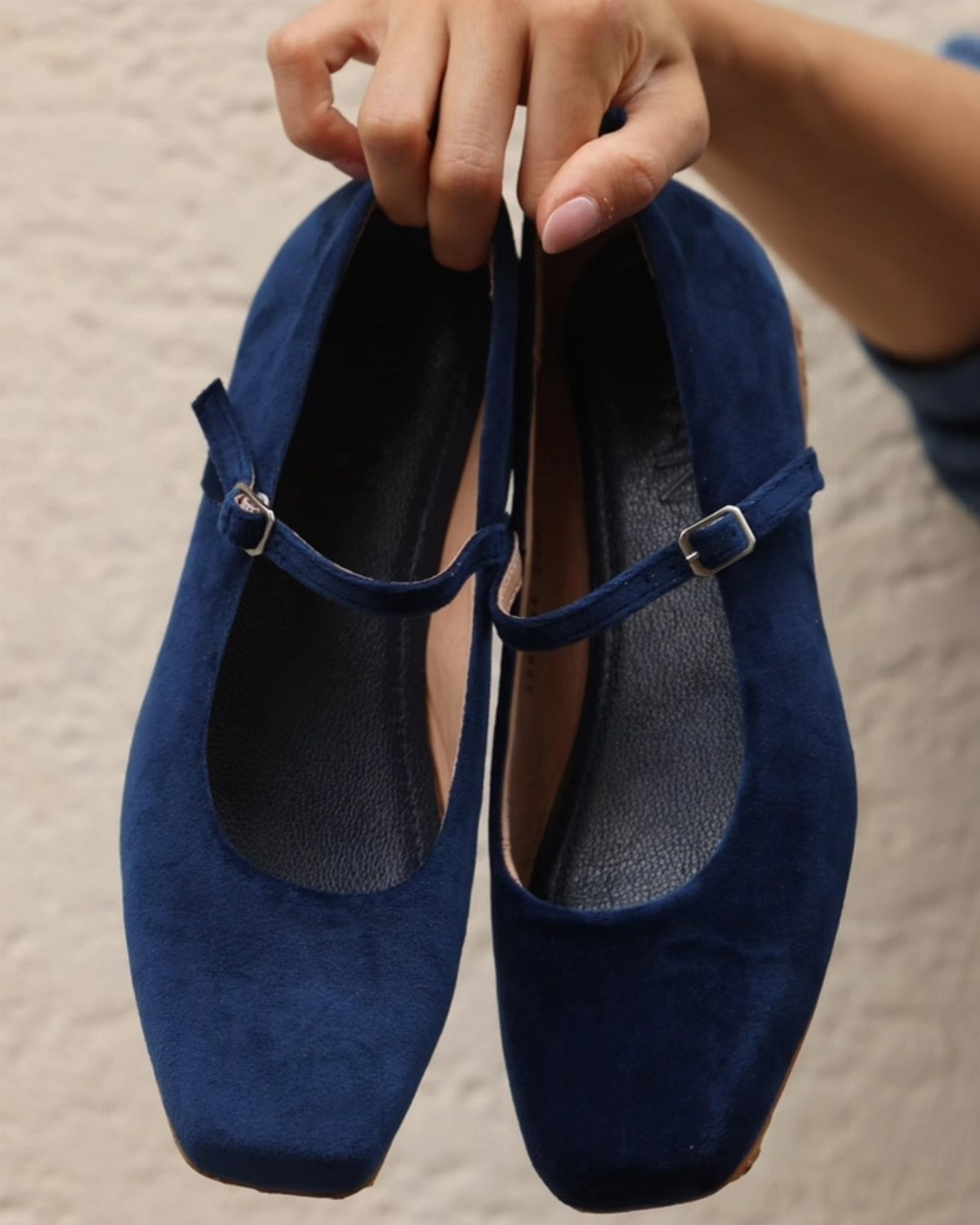 A hand holding a pair of navy blue velvet Mary Jane flats with silver buckles.
