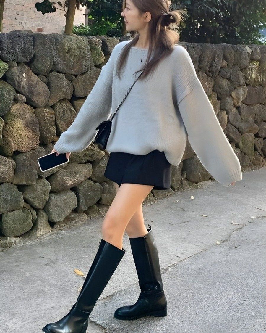 A woman wearing a grey sweater, black shorts, and tall black boots walking past a stone wall.