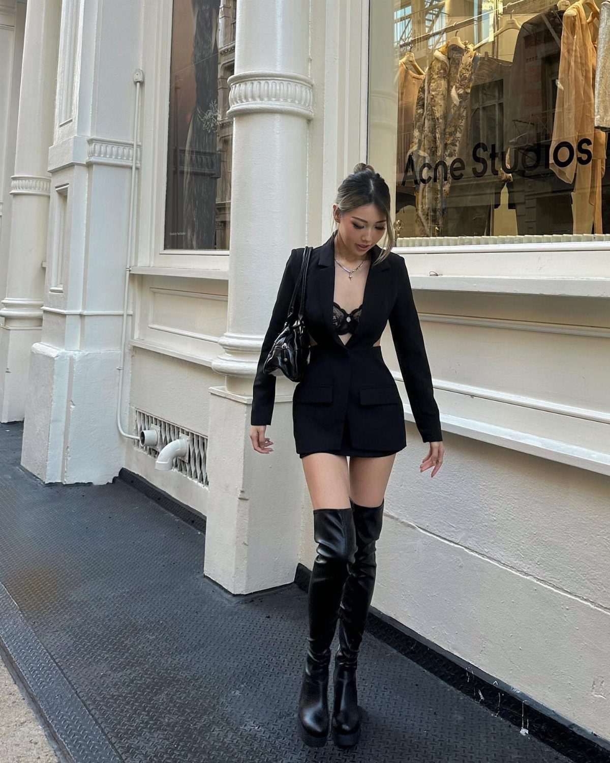 A woman in a black outfit and thigh-high boots walking past a white building.