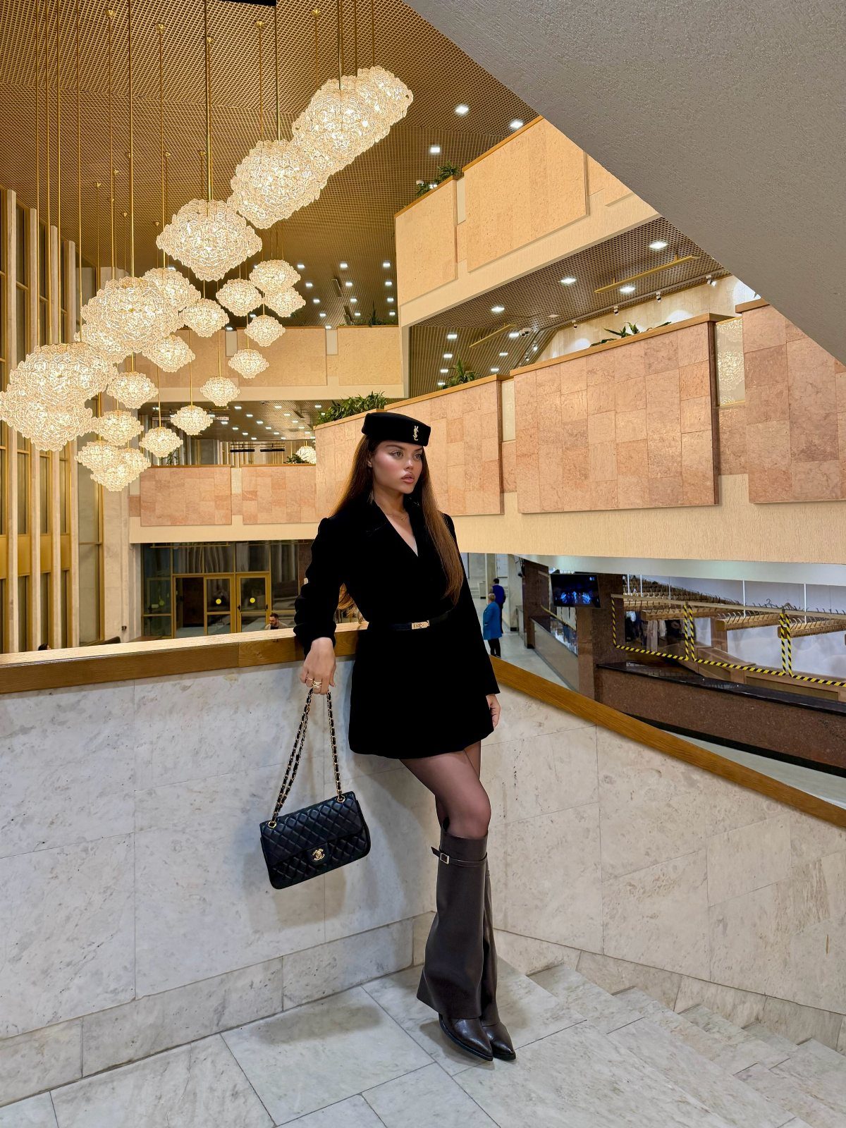 Woman wearing a black dress and tall black boots posing in a modern building.