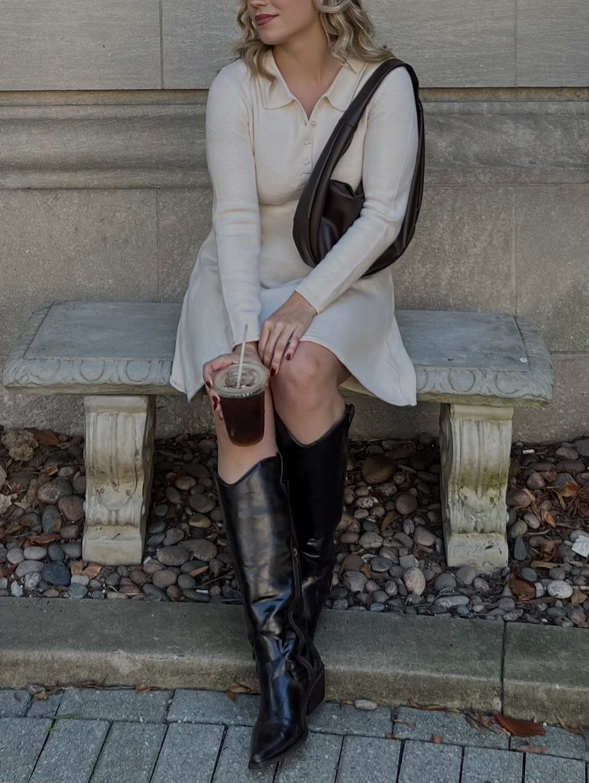 Woman sitting on a stone bench wearing a cream dress and tall black leather boots.
