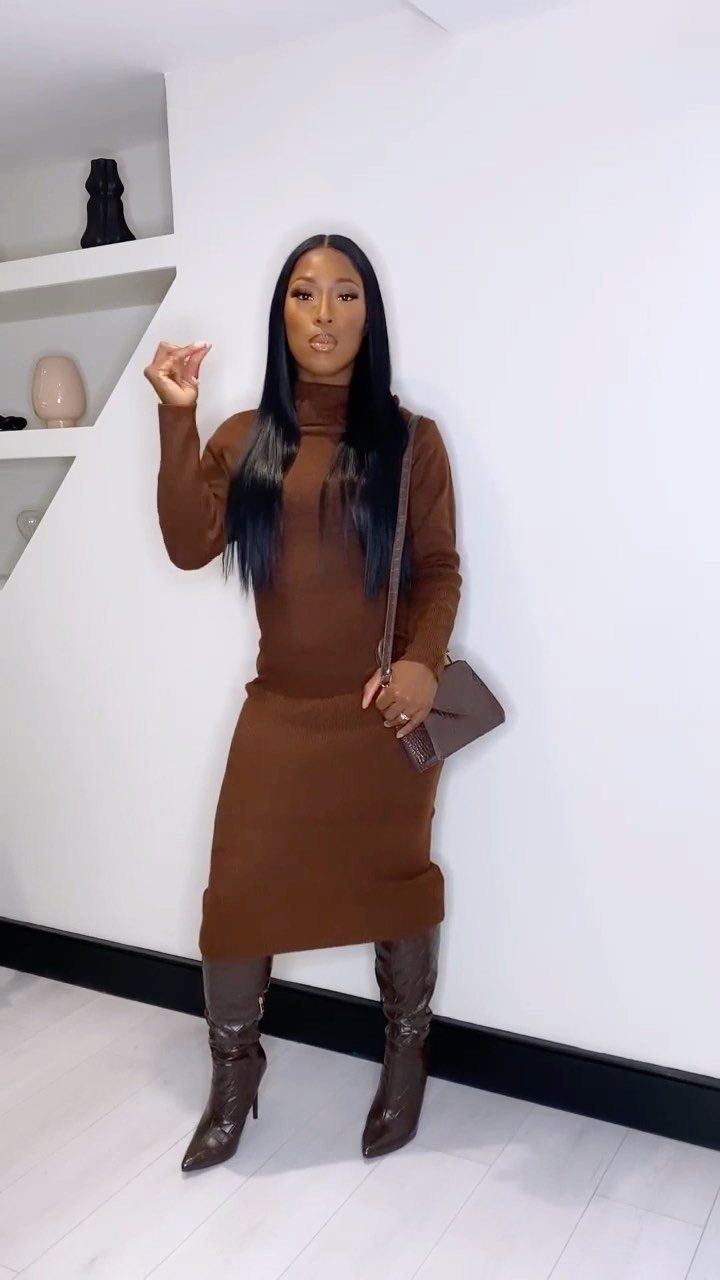 Woman in a brown turtleneck sweater dress and tall brown leather boots standing against a white wall.