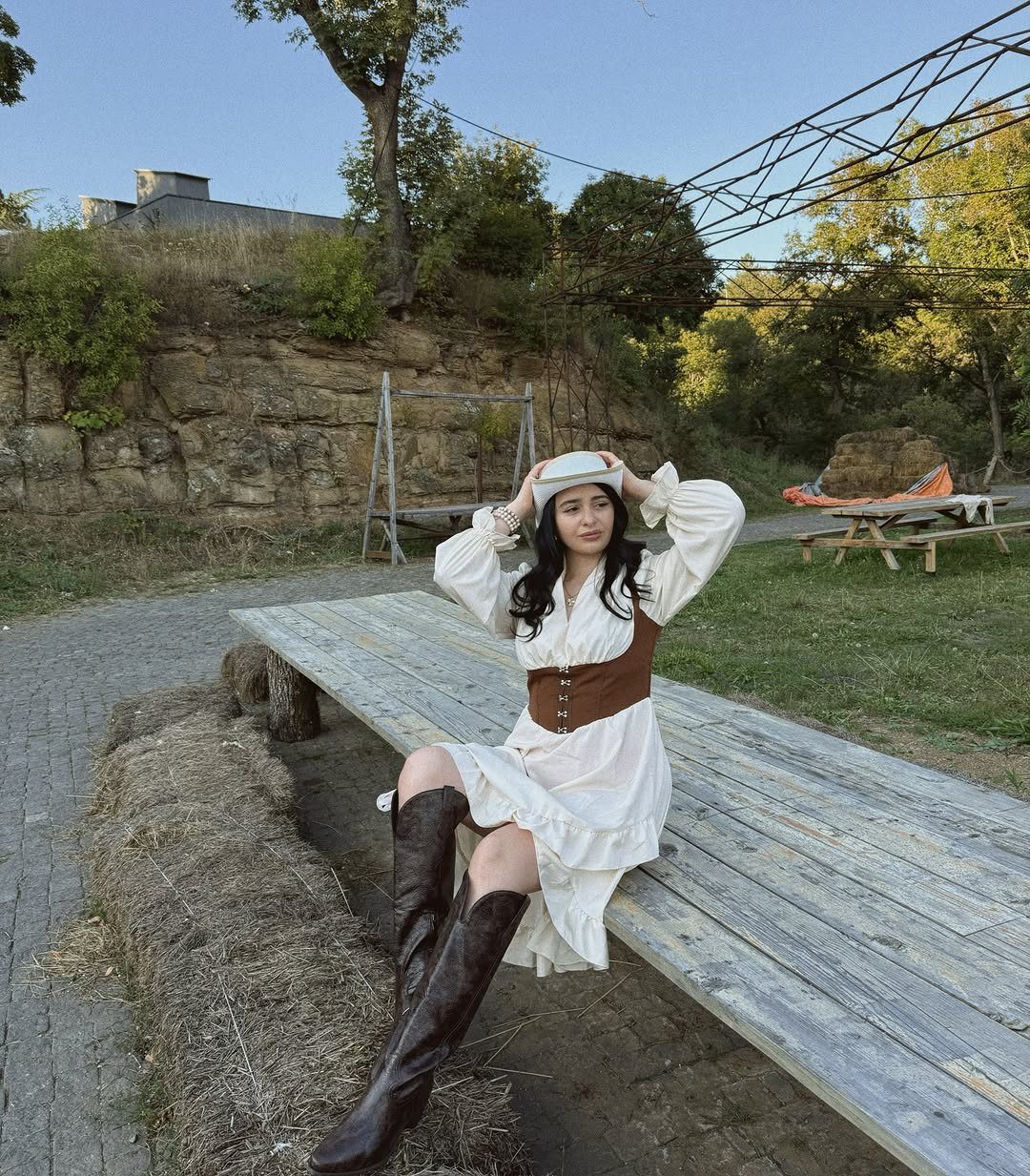 A woman in a white dress and brown leather boots sitting on a wooden bench outdoors.