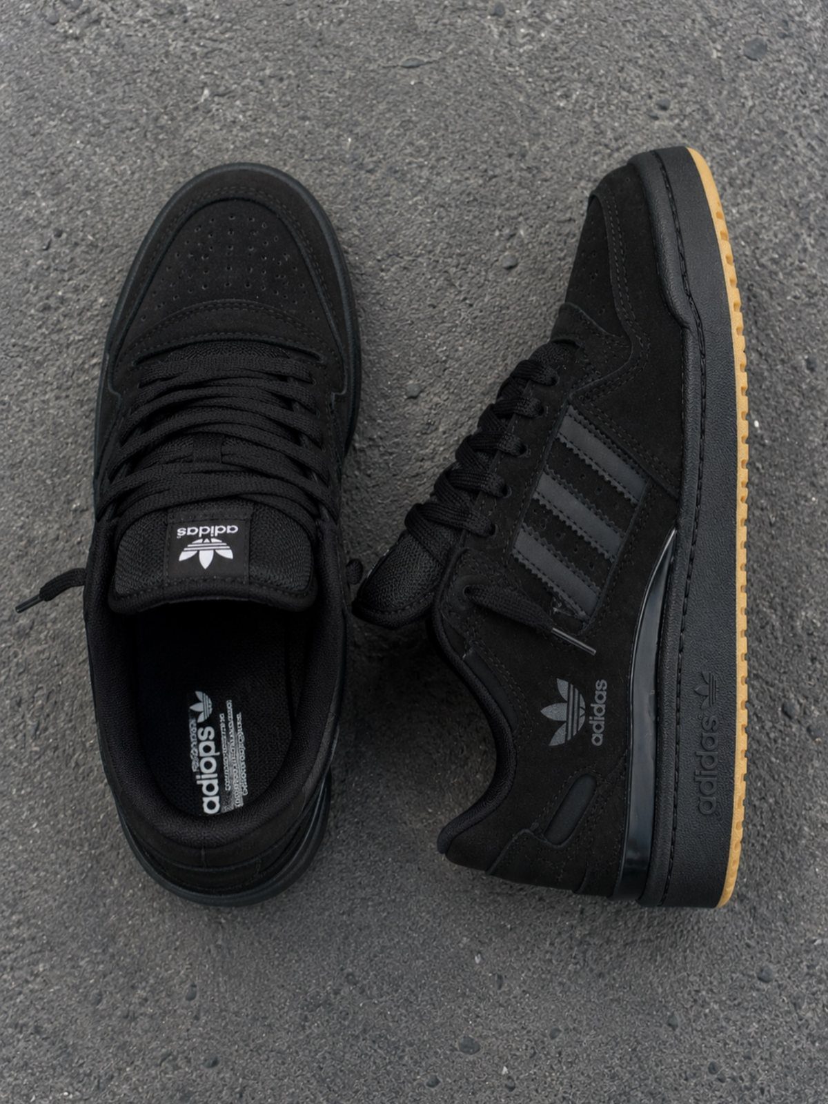 Two black Adidas sneakers with gum soles resting on a grey asphalt ground.