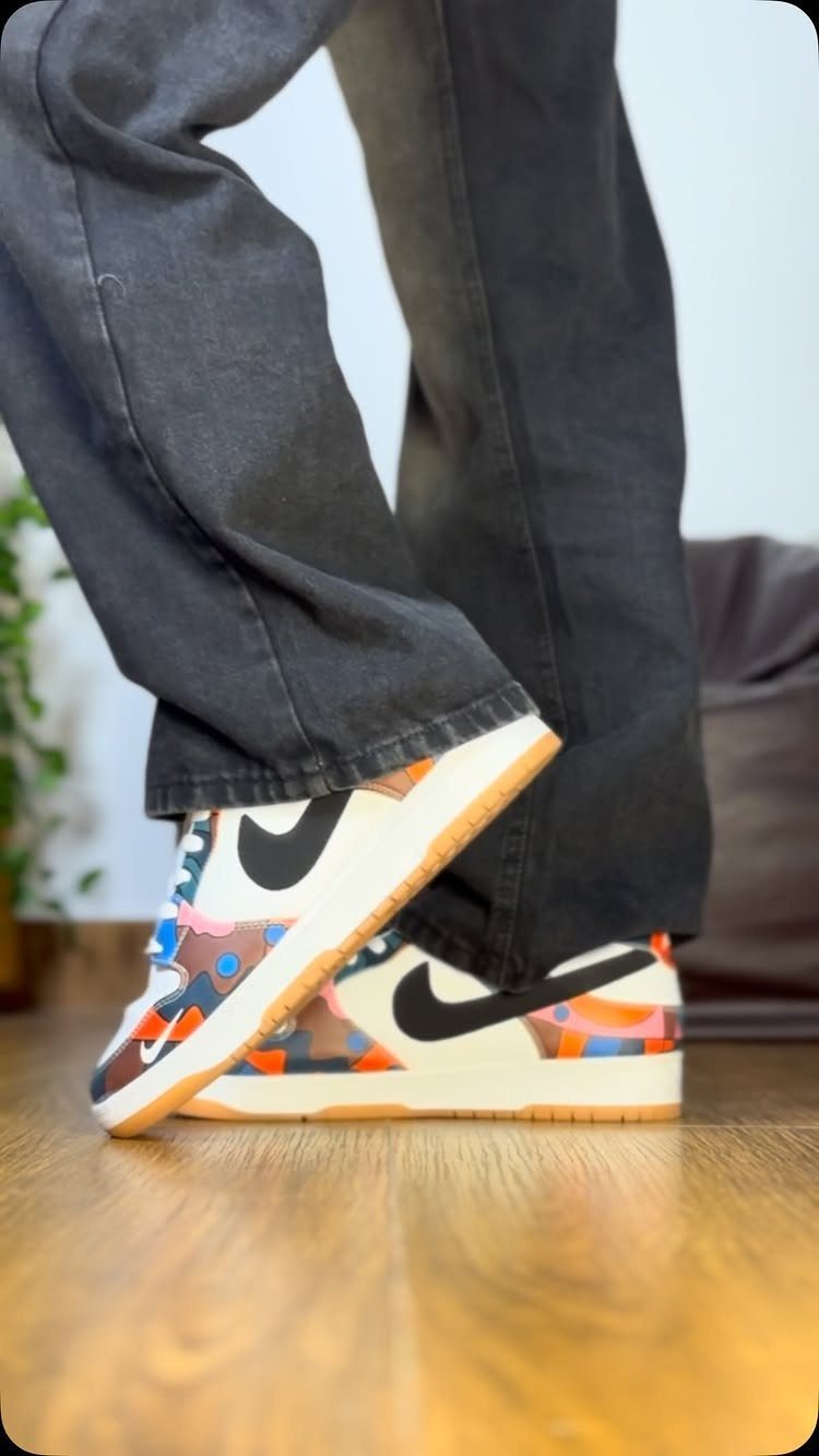 Person wearing dark grey wide-leg jeans and colorful patterned Nike sneakers on a wooden floor.