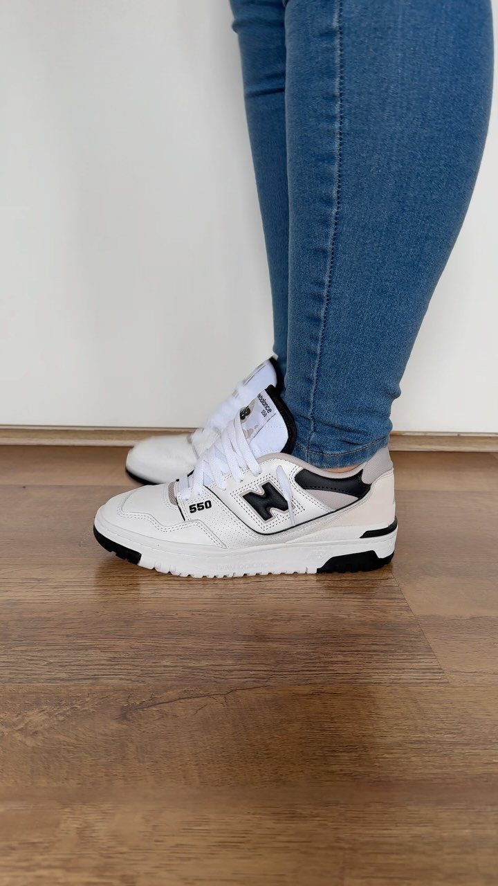 White New Balance 550 sneakers worn with blue jeans on a wooden floor.