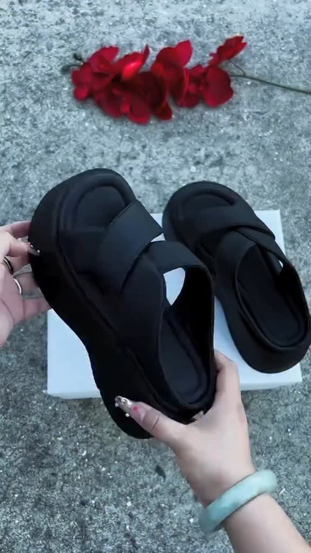 Hands holding a pair of chunky black platform slide sandals over a white box.