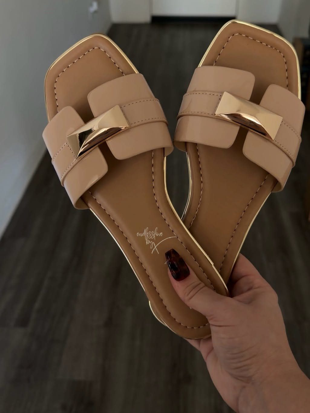 Tan flat open-toe slide sandals with wide straps and gold metallic buckles held by a hand.