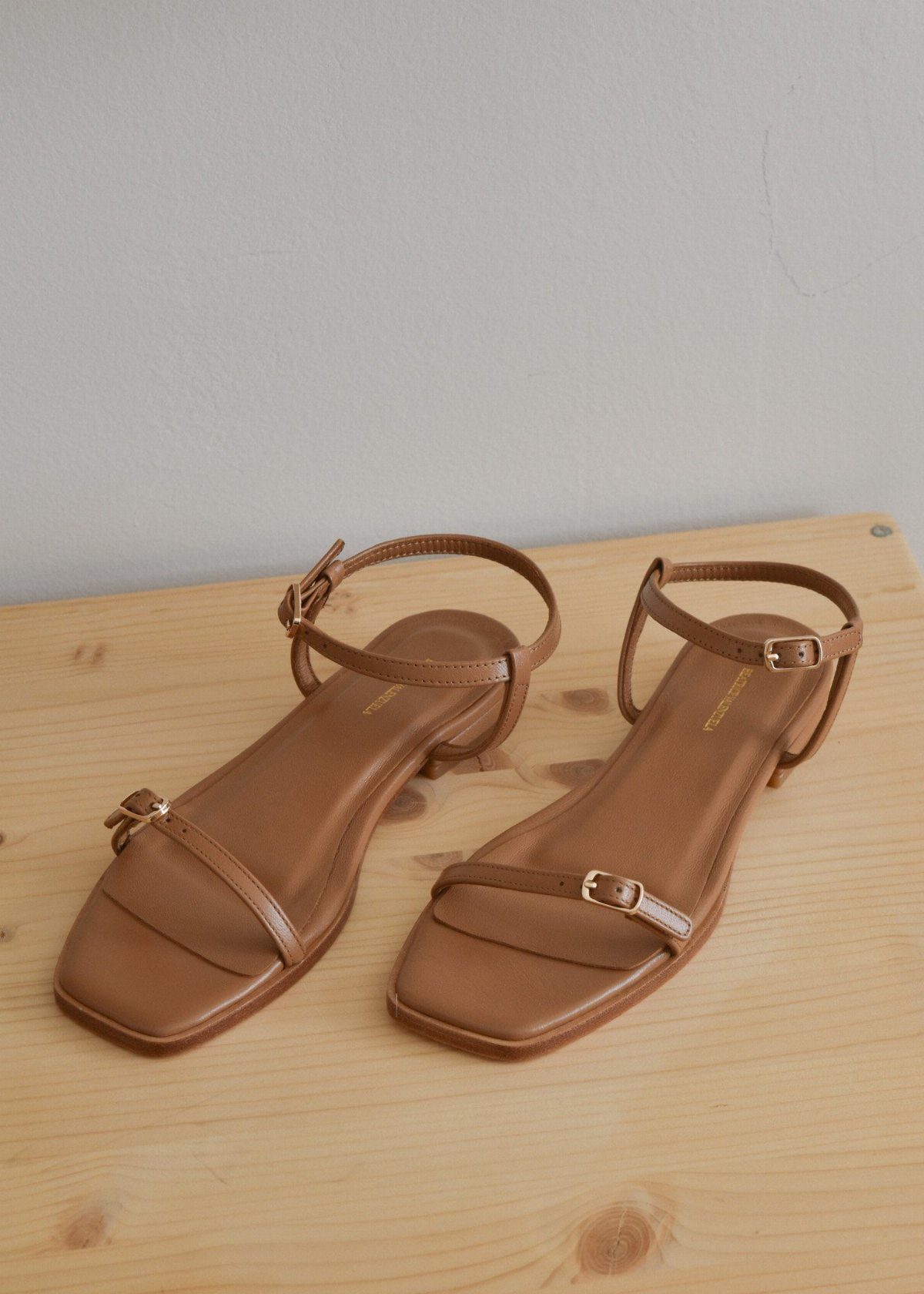 Tan leather flat sandals with square toes and thin ankle straps on a wooden table.
