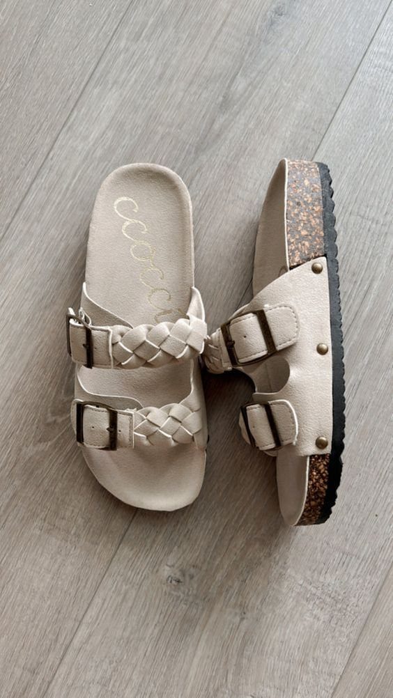 Beige braided strap slide sandals with metallic buckles resting on a light grey wooden floor.