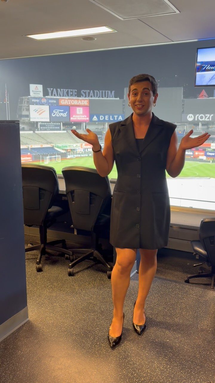 A woman in a black dress posing in an office with Yankee Stadium in the background.