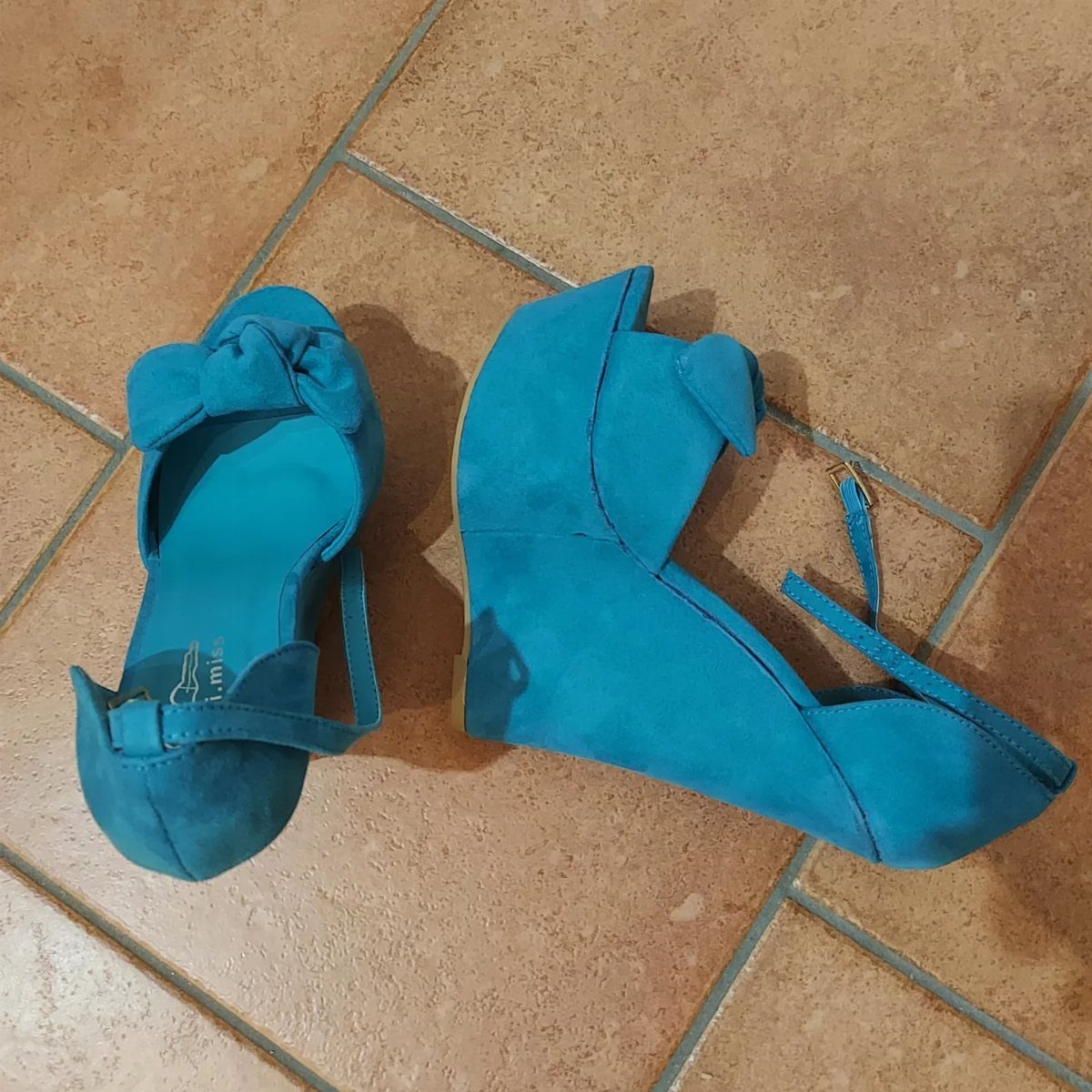 Turquoise blue suede wedge sandals with ankle straps and bows on a tan tile floor.