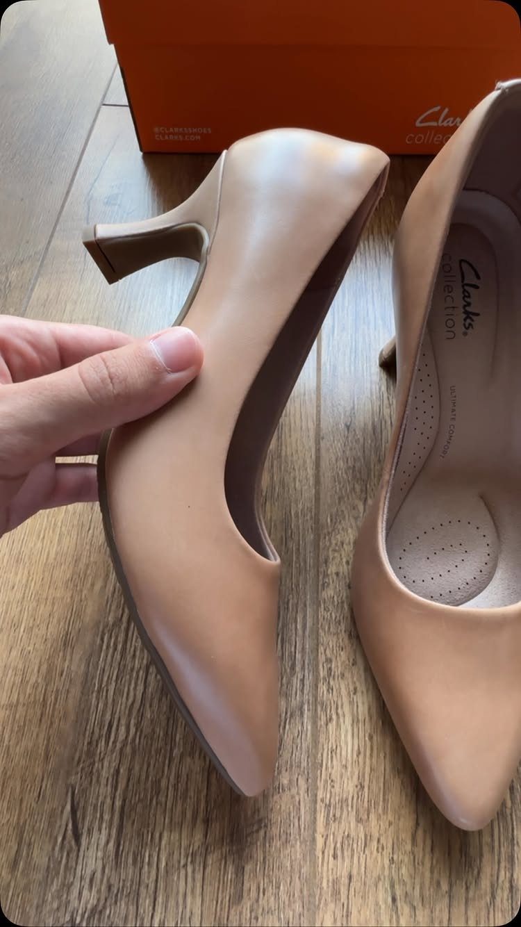 A hand holds a tan leather Clarks pump heel over a wooden floor.