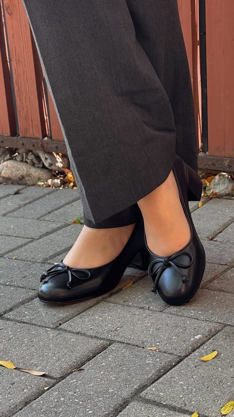Black leather ballet flats with bow details worn by a person on a brick walkway.
