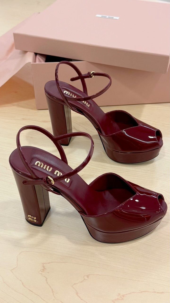 Burgundy patent leather platform high heels with ankle straps resting on a wooden surface near a pink box.