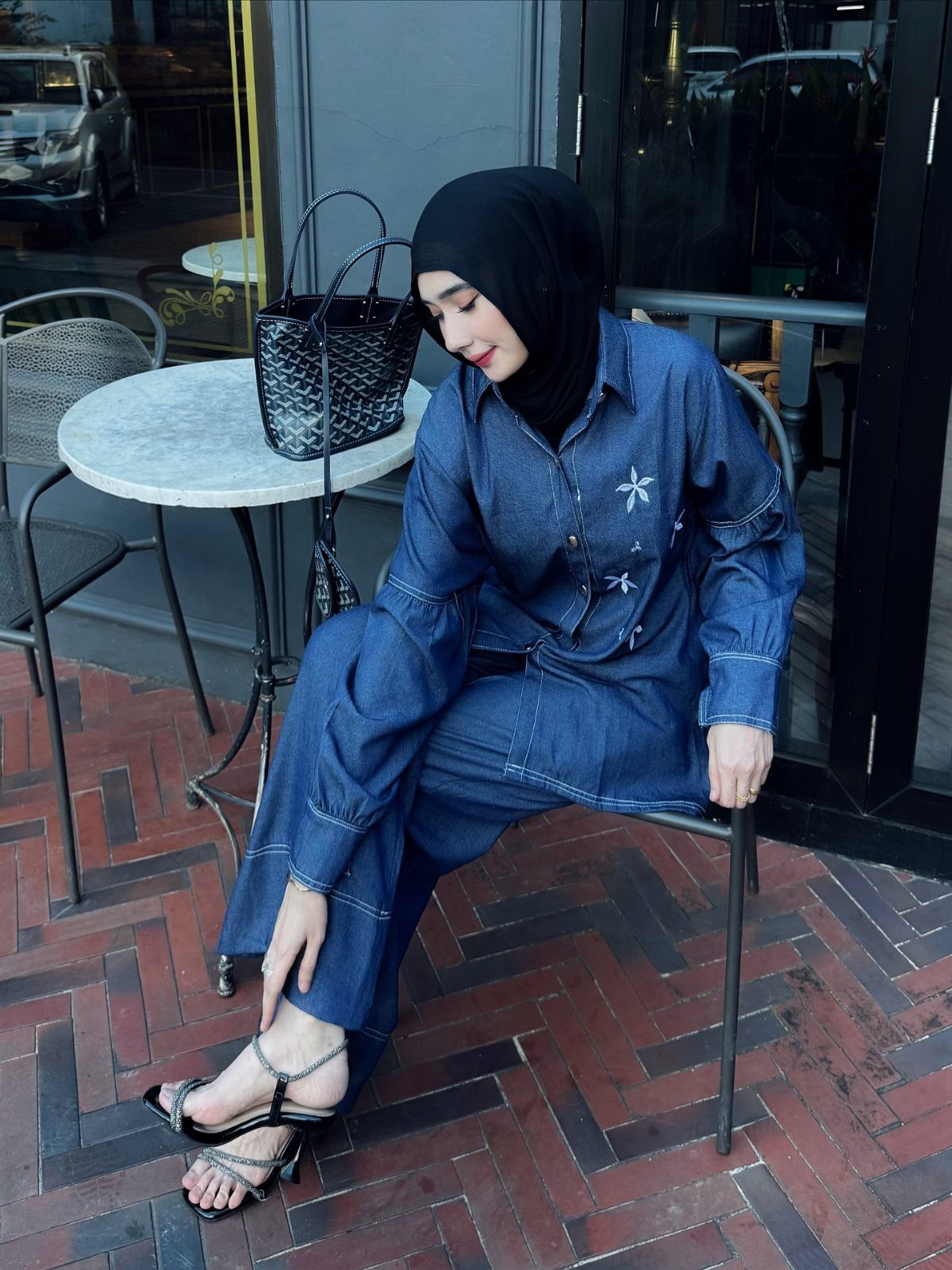 Woman in denim outfit and hijab sitting at a cafe wearing black strappy heeled sandals.