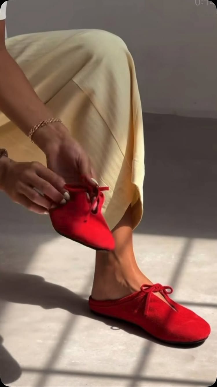 Red backless slide mules with bow details being held by a person wearing one.