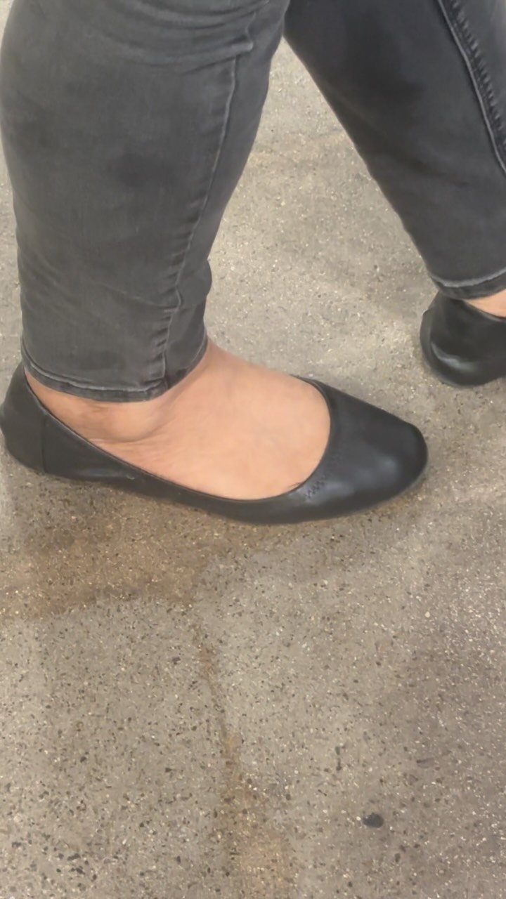 Black leather ballet flats worn by a person in grey jeans on concrete.