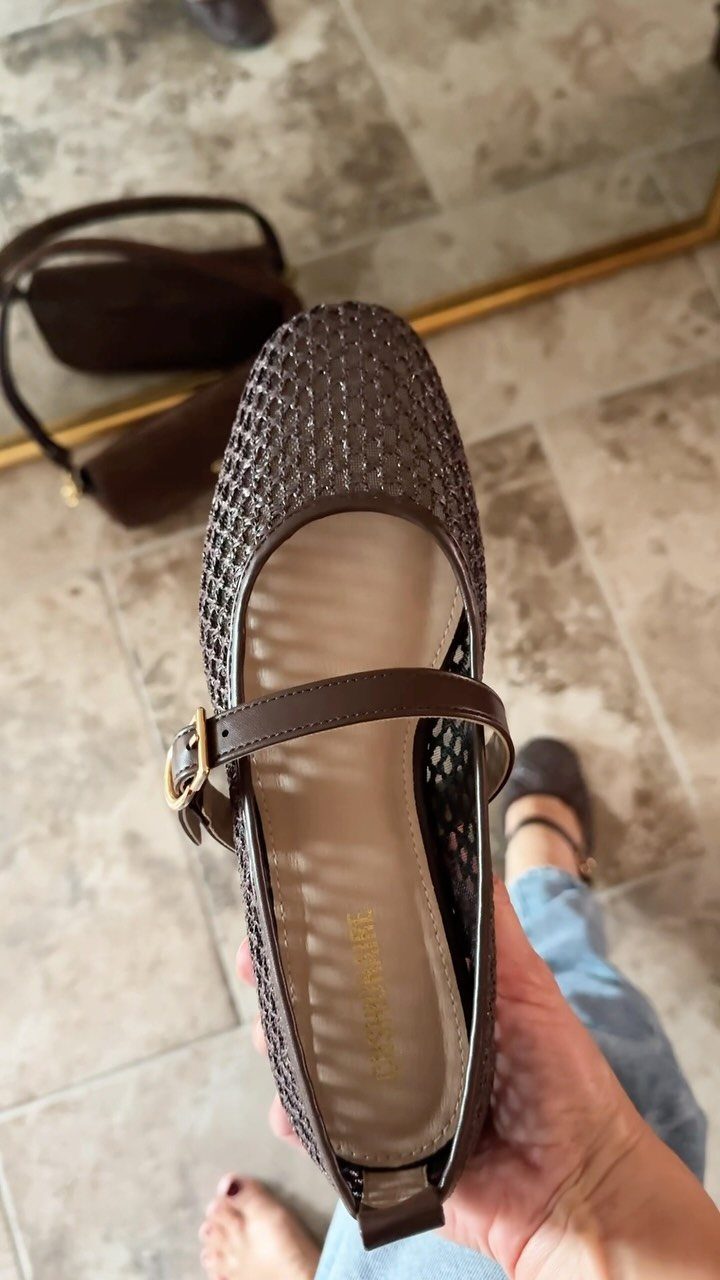A hand holding a brown woven Mary Jane flat shoe with a gold buckle strap.