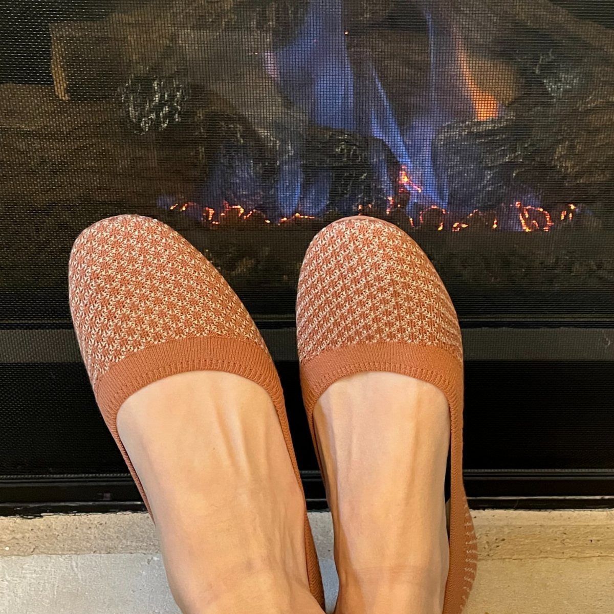 Feet wearing tan textured knit slip-on flats in front of a glowing fireplace.