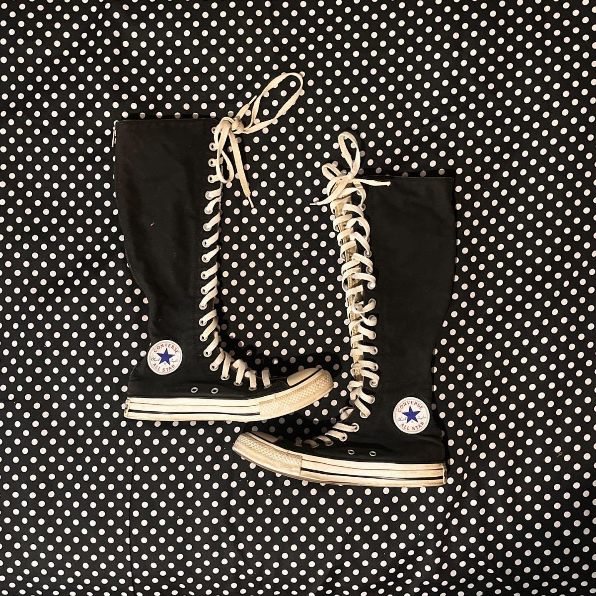 Black canvas knee-high sneakers with white laces resting on a black and white polka dot surface.