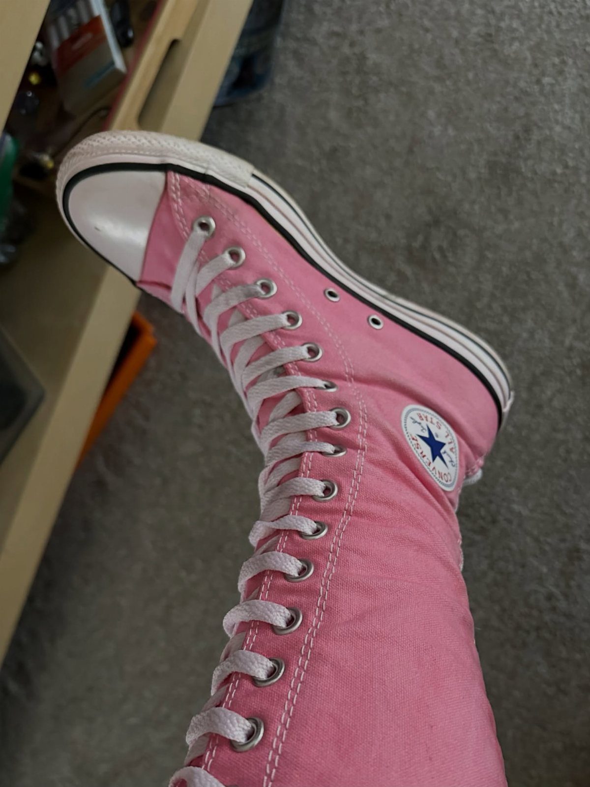 A pink knee-high Converse sneaker with white laces on a grey carpeted floor.