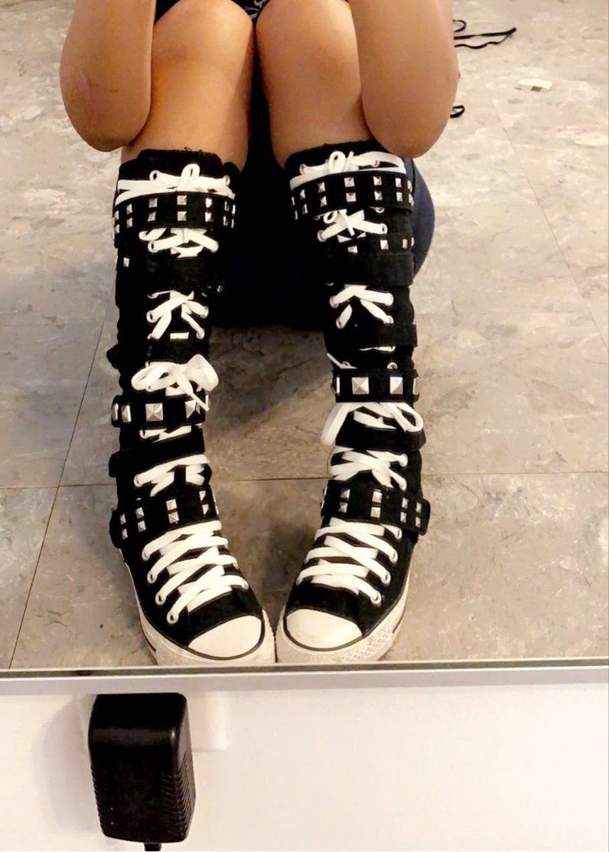 Black knee-high sneakers with white laces and silver studded straps worn by a person sitting down.