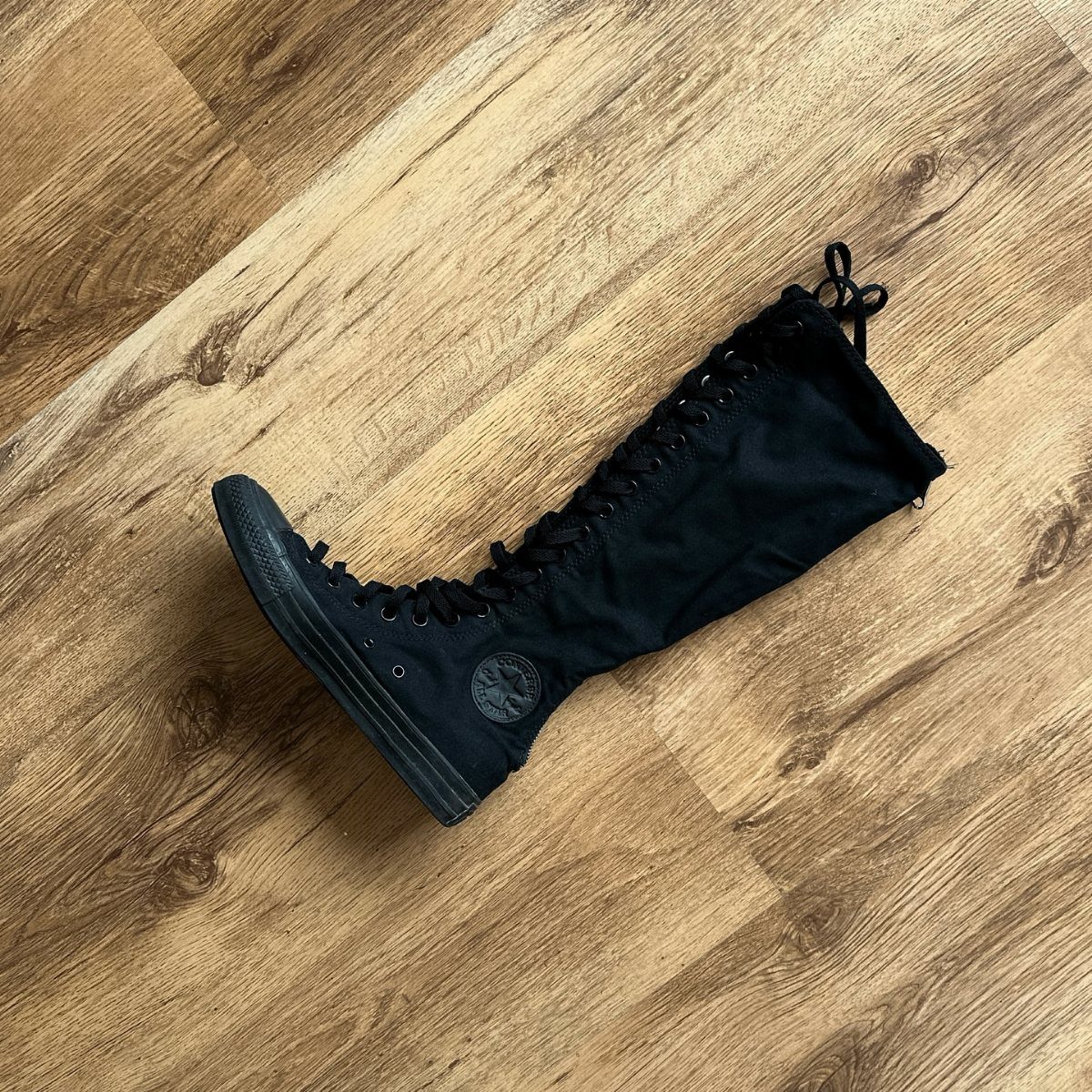 A single black canvas knee-high lace-up sneaker resting on a wooden floor.
