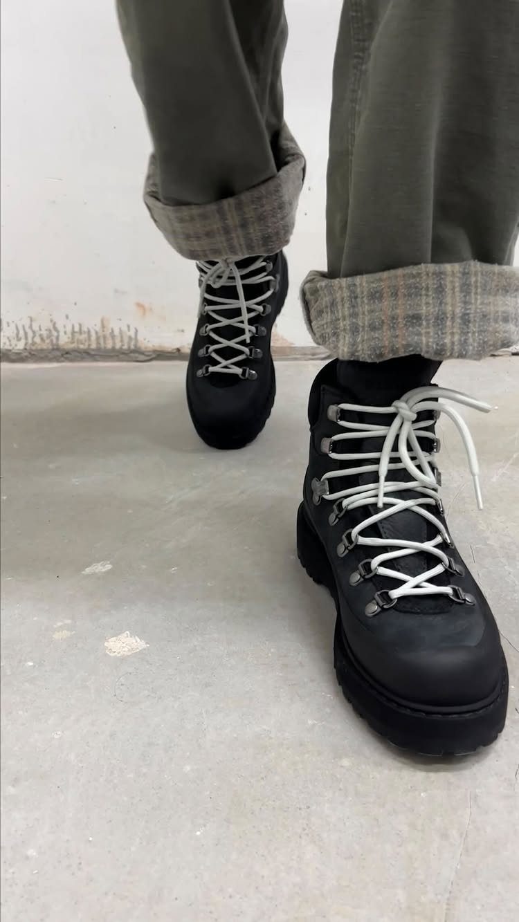 Black lace-up winter boots with white laces worn by a person in olive green trousers.