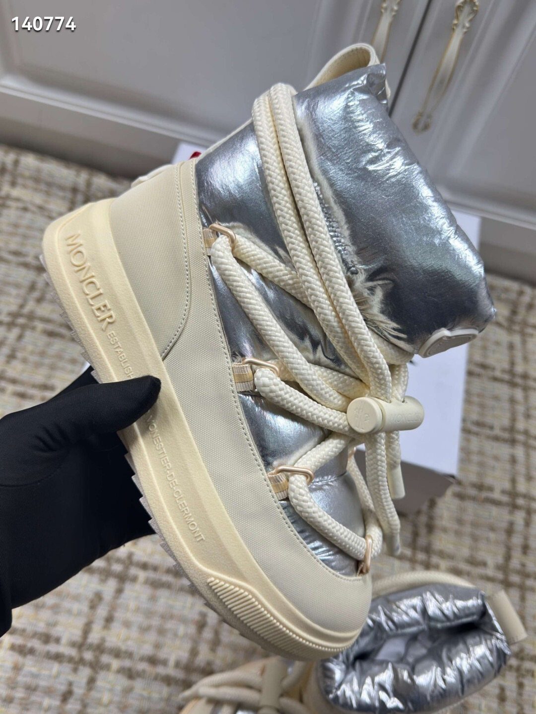 Silver metallic Moncler winter boot with thick cream laces held by a hand in a black glove.